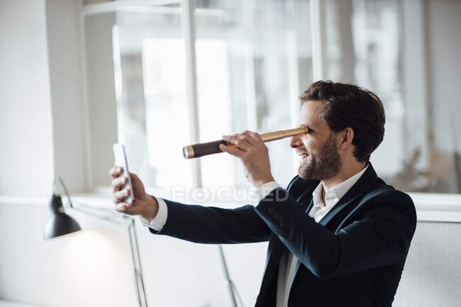 Businessman looking at mobile phone through binoculars in office — Stock Photo