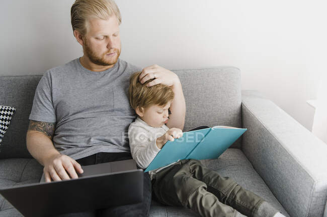 Сын читает книжку с картинками, а отец сидит дома с ноутбуком на диване — стоковое фото