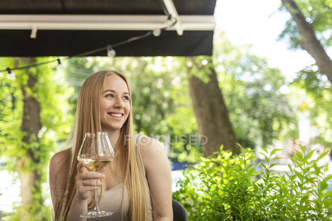 Sorridente giovane donna bionda seduta con bicchiere da vino al ristorante guardando altrove — Foto stock