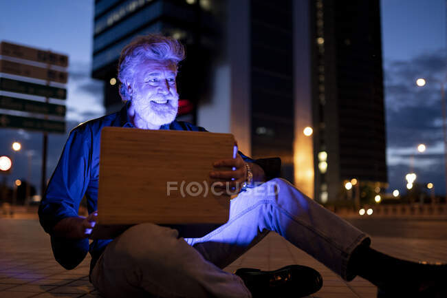 Empresário sorridente sentado com laptop no caminho da cidade à noite — Fotografia de Stock