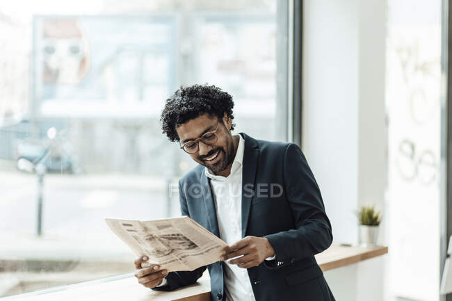 Hombre sonriente empresario leyendo el periódico mientras se inclina escritorio por ventana en la oficina - foto de stock