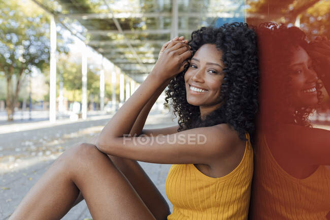 Mulher jovem sorridente bonita sentada na parede de vidro vermelho — Fotografia de Stock