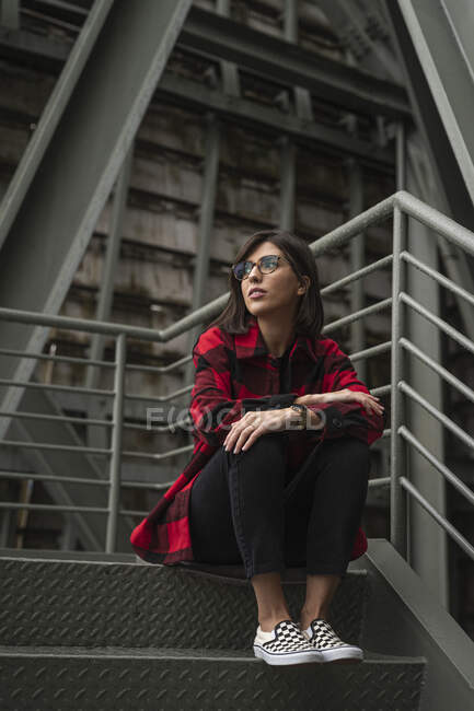 Jeune femme réfléchie avec des lunettes assis sur les marches par rambarde — Photo de stock