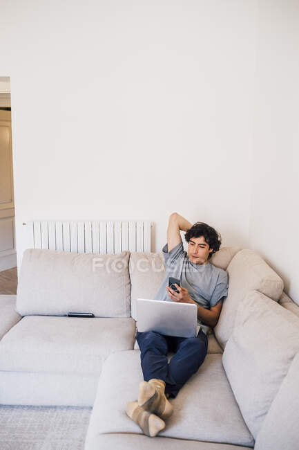 Uomo che utilizza il telefono cellulare mentre seduto con il computer portatile sul divano in soggiorno — Foto stock