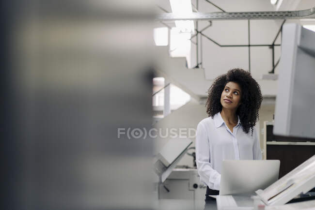 Femme d'affaires avec ordinateur portable regardant loin tout en se tenant à la machinerie dans l'industrie — Photo de stock