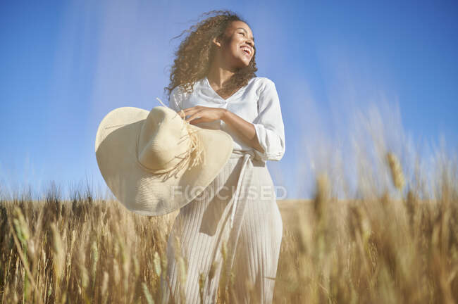 Joyeux jeune femme avec chapeau debout dans le champ de blé tout en regardant loin — Photo de stock