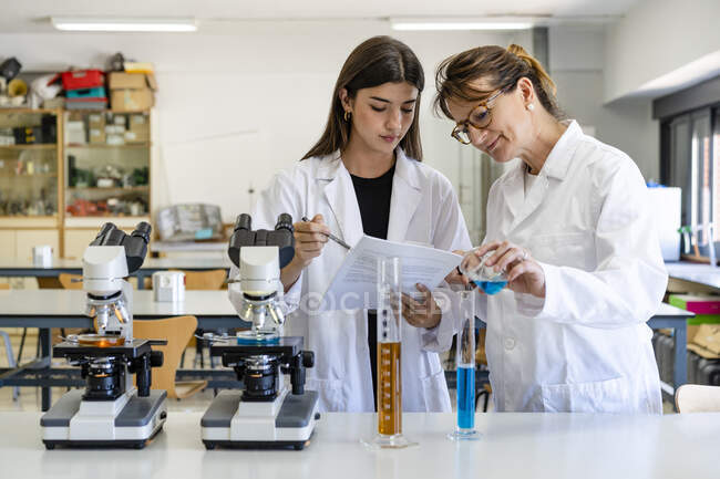 Female scientist reading document while colleague pouring liquid in graduated cylinder — Stock Photo