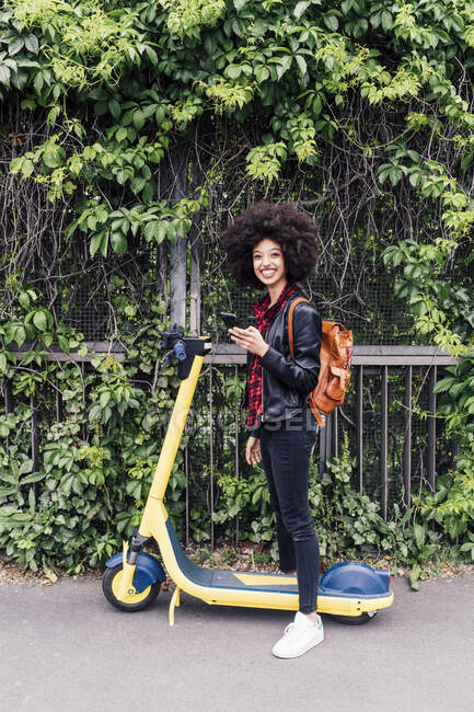 Femme souriante tenant téléphone portable tout en se tenant debout avec scooter électrique push sur la route — Photo de stock