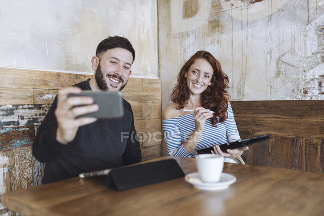 Homme prenant selfie avec une collègue femme tenant tablette graphique au café — Photo de stock