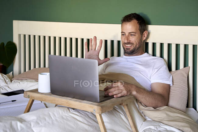 Uomo sorridente che agita mano a mano la videochiamata attraverso il computer portatile a casa — Foto stock
