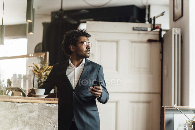Imprenditore maschio in piedi con smart phone e tazza di caffè in caffè guardando altrove — Foto stock