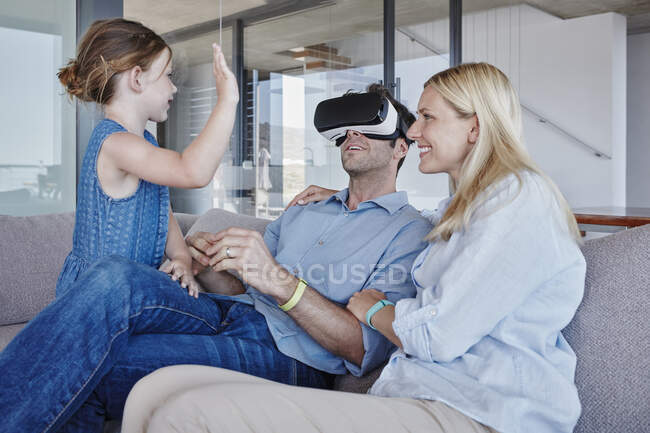Девушка машет отцу в симуляторе виртуальной реальности, сидя рядом с женщиной в гостиной — стоковое фото
