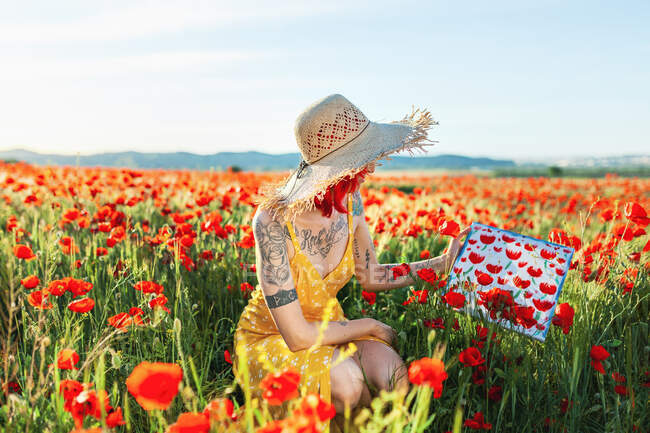 Mulher tatuada segurando pintura em meio a flores de papoula no campo — Fotografia de Stock
