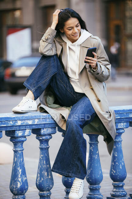 Donna sorridente utilizzando il telefono cellulare mentre seduto su ringhiera blu — Foto stock