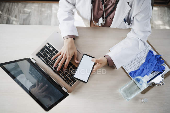 Médico femenino sosteniendo el teléfono inteligente mientras usa el portátil en el escritorio - foto de stock