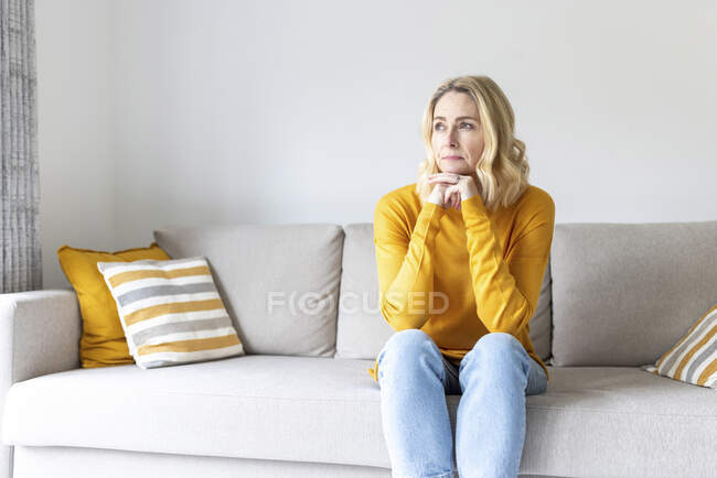 Mulher cuidadosa com a mão no queixo sentado no sofá em casa — Fotografia de Stock