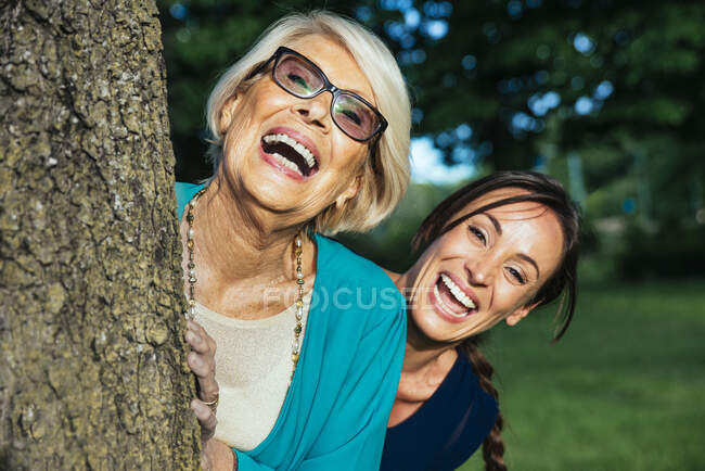 Donna mezza adulta spensierata con donna anziana che sbircia dall'albero nel parco pubblico — Foto stock