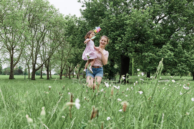 Mère heureuse portant fille avec jouet pinwheel tout en marchant sur l'herbe au parc — Photo de stock