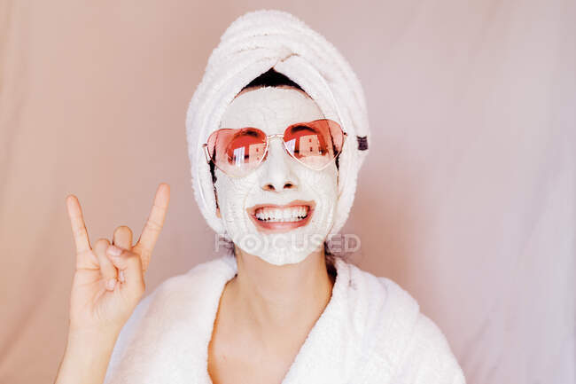 Jovem feliz com máscara facial de beleza mostrando sinal de chifre contra fundo de pêssego — Fotografia de Stock