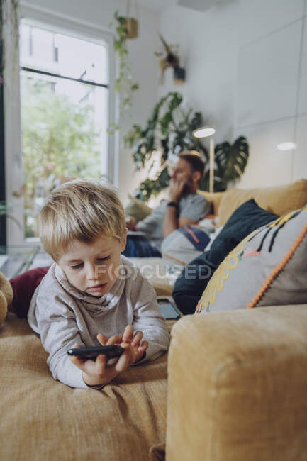 Мальчик пользуется мобильным телефоном, лежа на диване в гостиной — стоковое фото