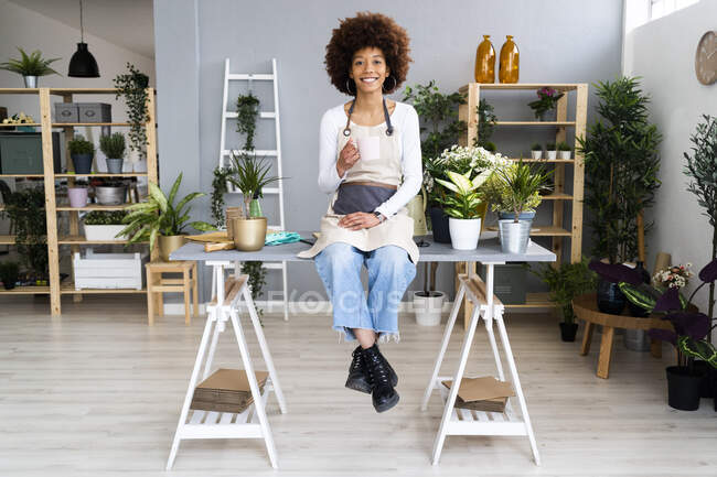 Femme propriétaire d'un magasin tenant un café tout en étant assis sur la table dans un magasin de plantes — Photo de stock