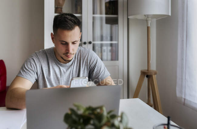 Junge männliche Unternehmer verwenden Smartphone, während sie mit Laptop zu Hause im Büro sitzen — Stockfoto