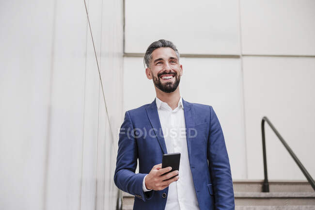 Männlicher Unternehmer mit Smartphone lächelt an der Wand — Stockfoto