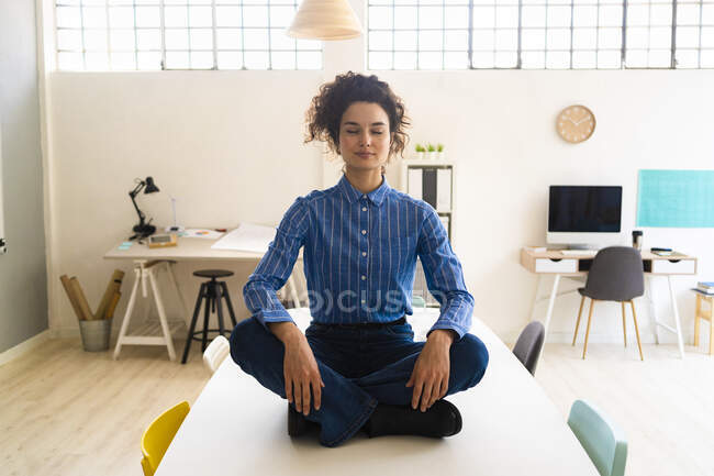 Mujer de negocios practicando yoga mientras está sentada con las piernas cruzadas en el escritorio en la oficina - foto de stock