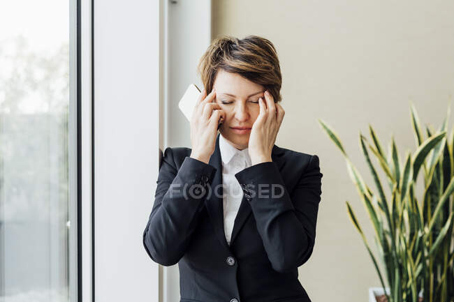 Уставшая деловая женщина с закрытыми глазами держит мобильный телефон в офисе — стоковое фото
