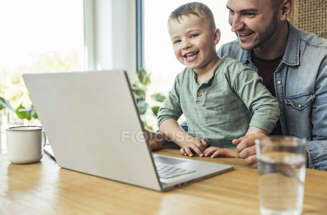 Веселые сын и отец сидят с ноутбуком делая видеозвонок на дому — стоковое фото
