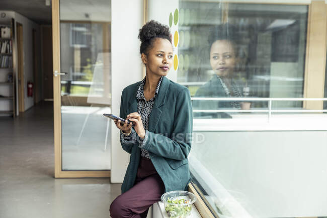 Female entrepreneur with mobile phone looking away while sitting on window sill at office — Stock Photo