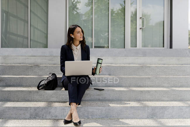 Femme d'affaires avec ordinateur portable regardant loin tenant tasse de voyage tout en étant assis sur l'escalier — Photo de stock