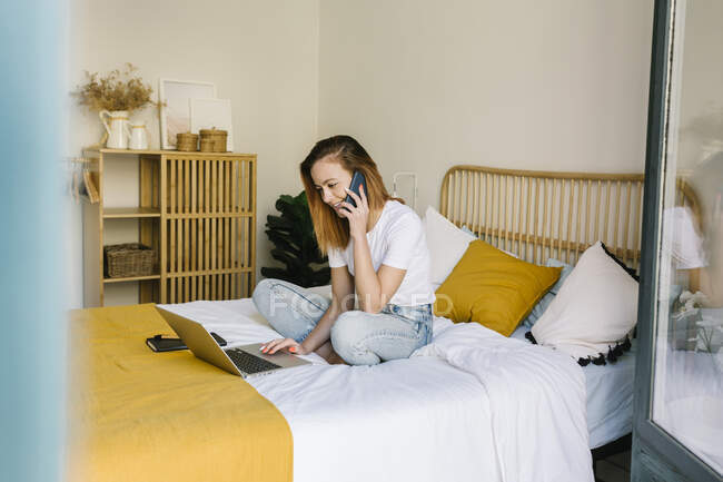 Улыбающаяся женщина разговаривает по мобильному телефону, сидя дома на кровати — стоковое фото