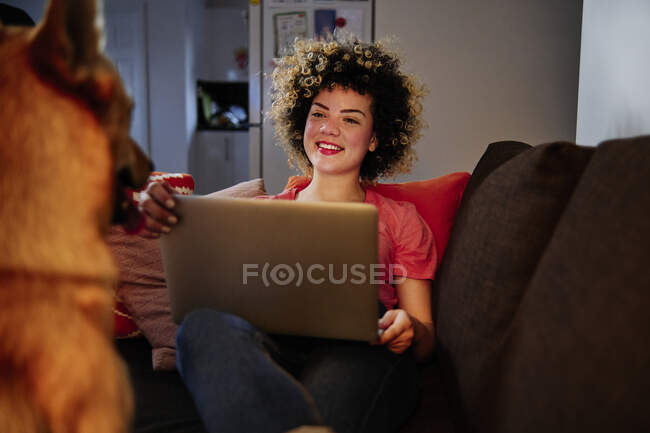 Sorridente giovane donna seduta con computer portatile guardando il cane in soggiorno — Foto stock