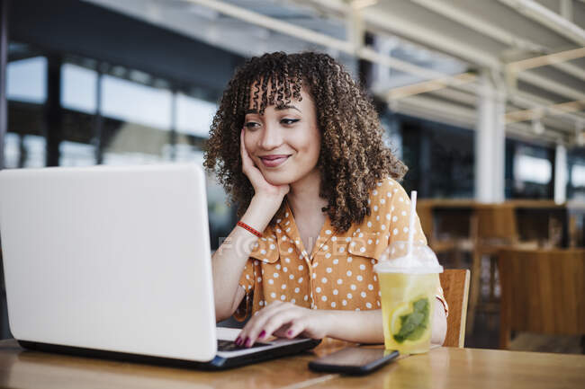 Smiling businesswoman with hand on chin using laptop at coffee shop — Stock Photo