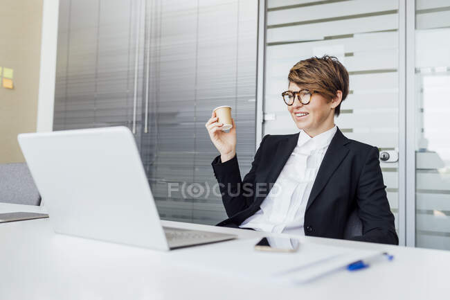 Smiling businesswoman having coffee while looking at laptop at desk — Stock Photo