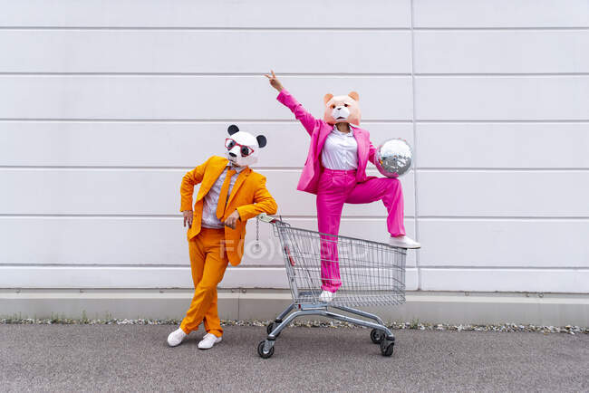 Man and woman wearing vibrant suits and animal masks posing with shopping cart and disco ball in front of white wall — Stock Photo