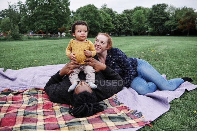 Familia feliz relajándose con su hijo mientras está acostado en la manta de picnic en el parque - foto de stock
