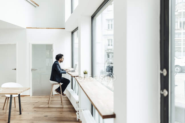Reifer Geschäftsmann arbeitet am Laptop, während er im Büro am Fenster sitzt — Stockfoto