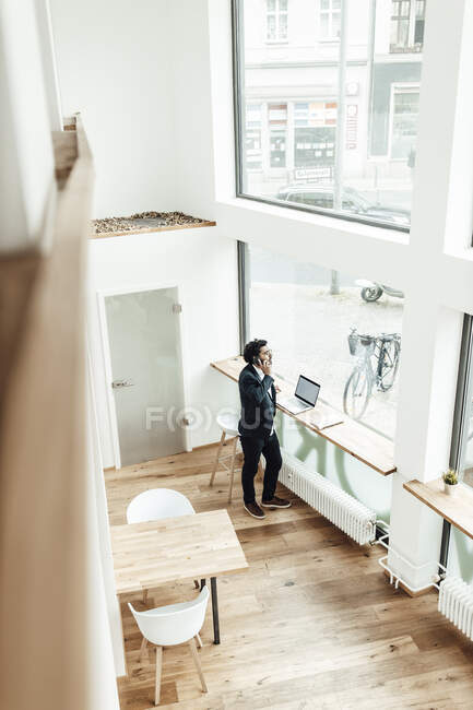 Reifer Geschäftsmann spricht auf Smartphone, während er im Büro durchs Fenster schaut — Stockfoto