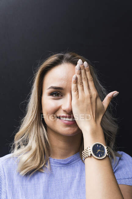 Souriant jeune femme couvrant l'oeil avec la main devant le mur noir — Photo de stock