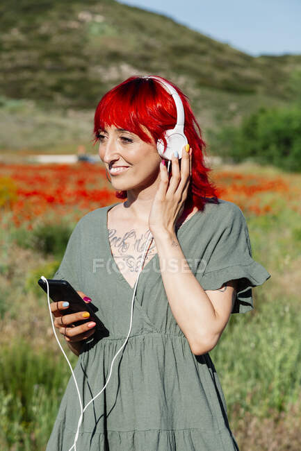 Улыбающаяся молодая женщина с мобильным телефоном слушает музыку через наушники на маковом поле — стоковое фото