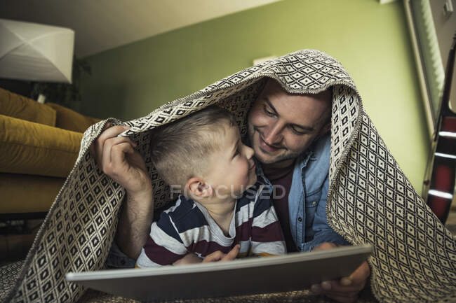 Sonrientes padre e hijo mirándose mientras están bajo la manta en la sala de estar en casa - foto de stock