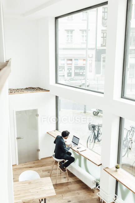 Männlicher Profi benutzt Laptop, während er im Büro am Glasfenster sitzt — Stockfoto