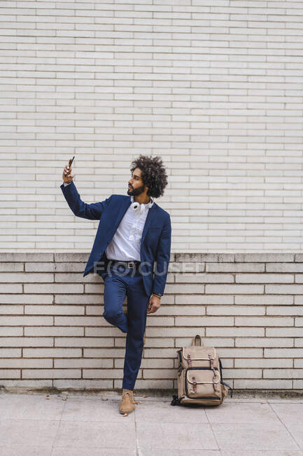 Businessman taking selfie through mobile phone while standing by backpack on footpath — Stock Photo