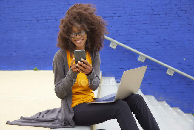 Femme souriante utilisant un téléphone mobile tout en étant assis avec un ordinateur portable sur les marches — Photo de stock