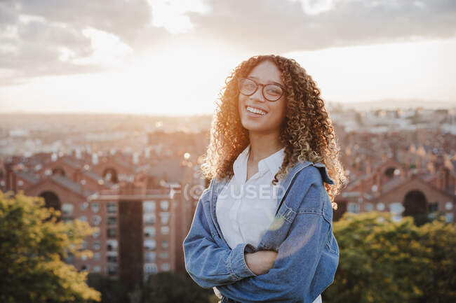Happy woman with arms crossed in front of city during sunset — Stock Photo