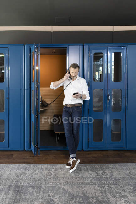Businessman using mobile phone while talking on telephone at booth in office — Stock Photo