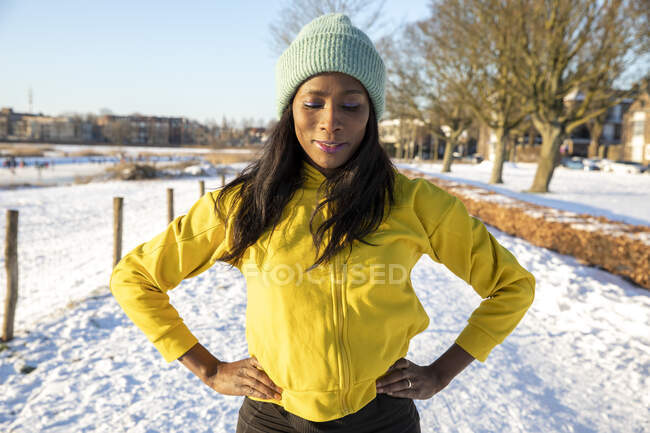 Усміхнена жінка в трикотажному капелюсі стоїть руками на стегні взимку — стокове фото