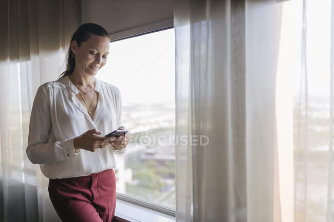 Female professional text messaging through mobile phone while standing by window in office — Stock Photo